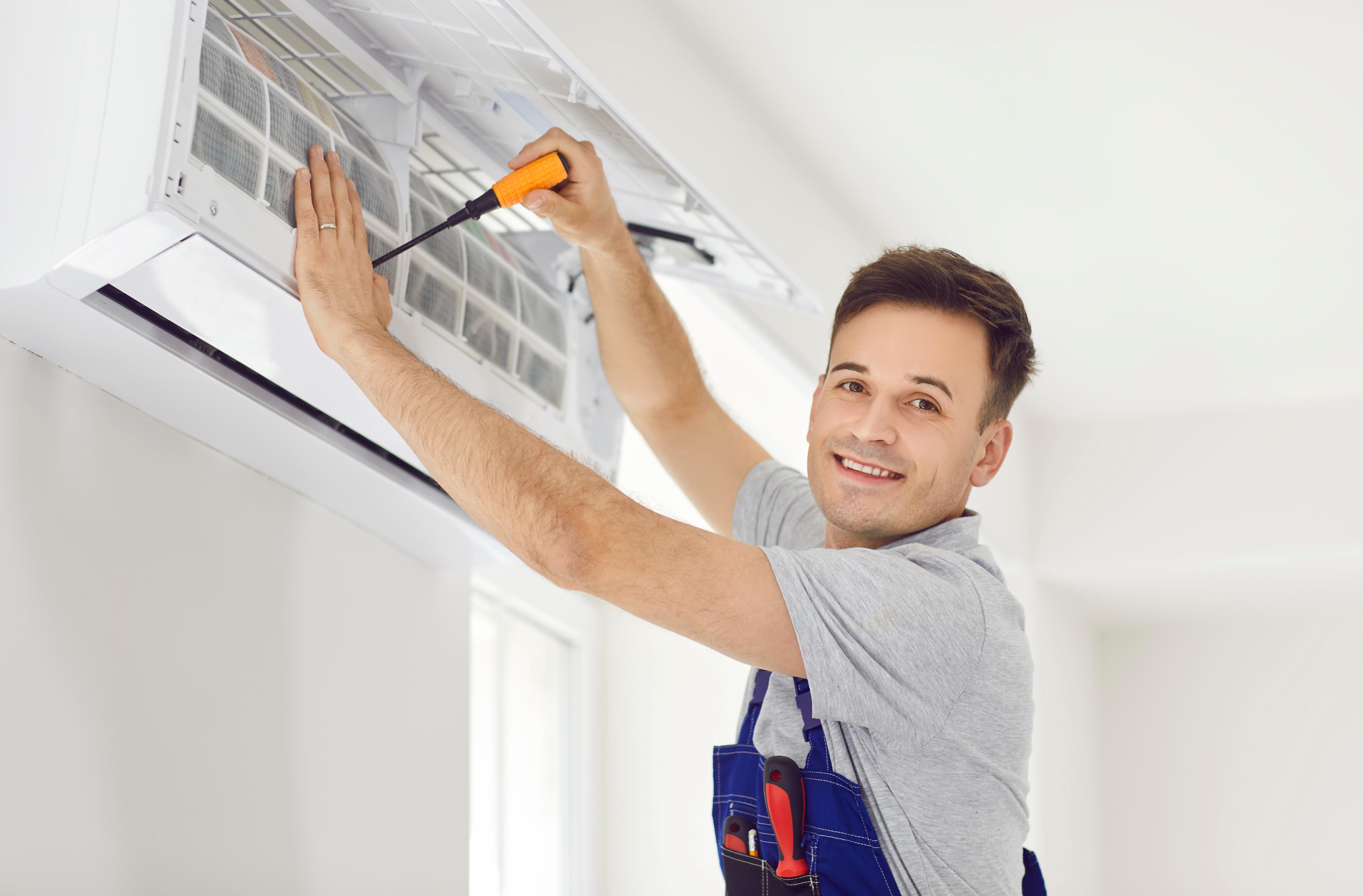 A smiling technician in overalls repairs an air conditioner with a screwdriver. The room is bright, conveying a positive and professional atmosphere.