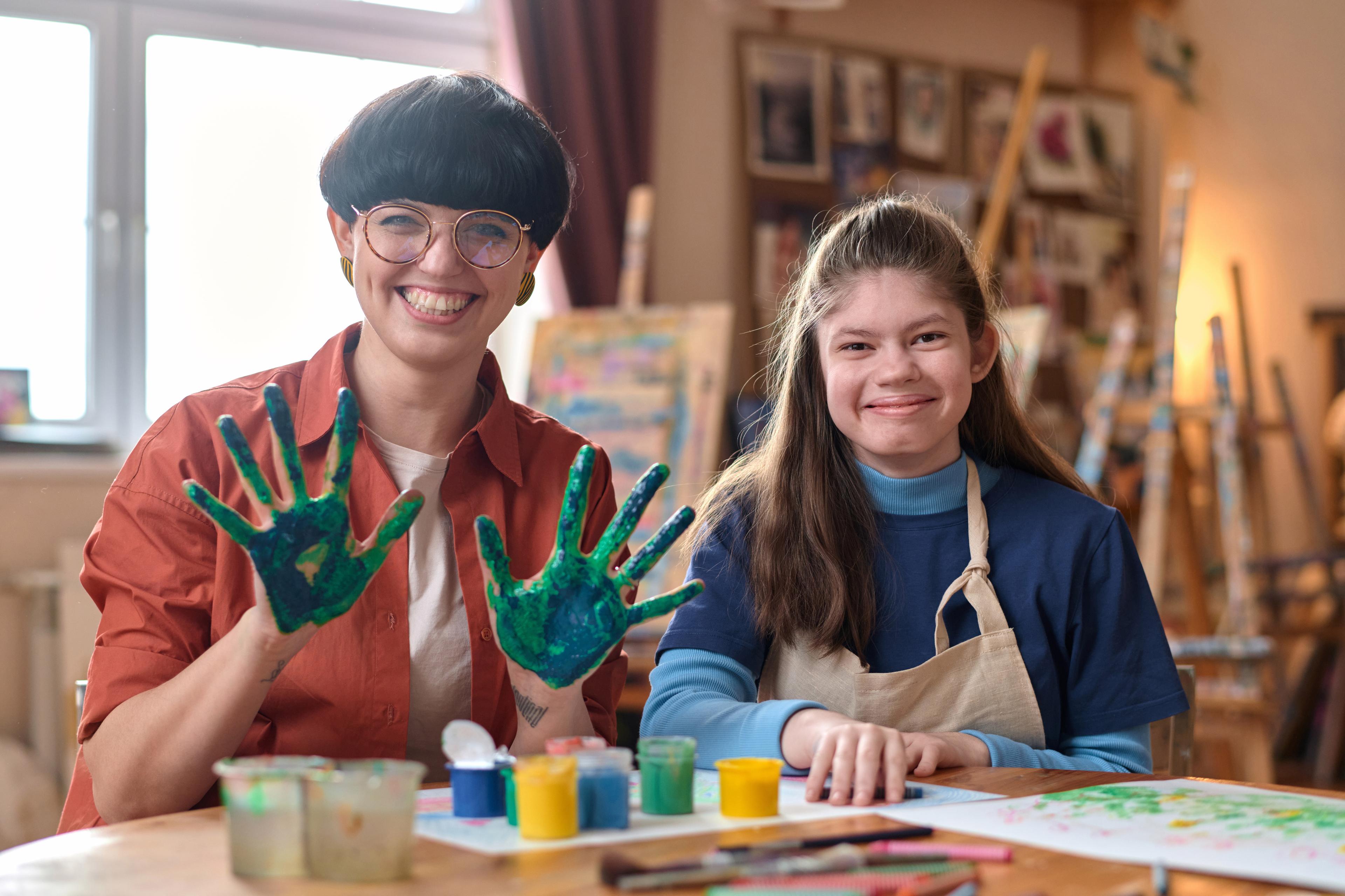 Two people in an art studio, smiling warmly. One shows paint-covered hands while the other sits beside paint jars and a colorful painting. Bright and cheerful atmosphere.