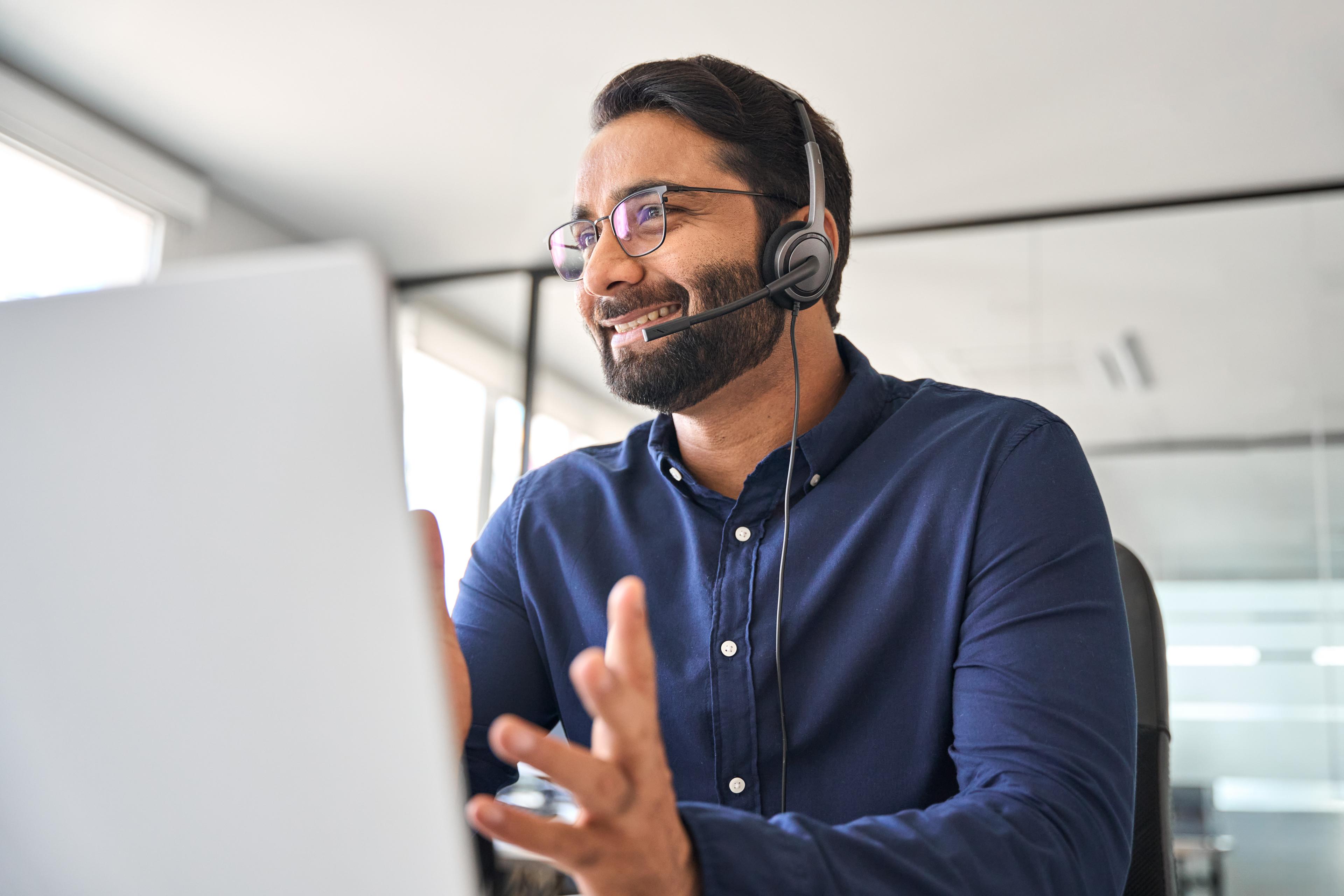 A man wearing glasses and a headset smiles while gesturing during a video call in a bright office setting, conveying a professional and positive mood.