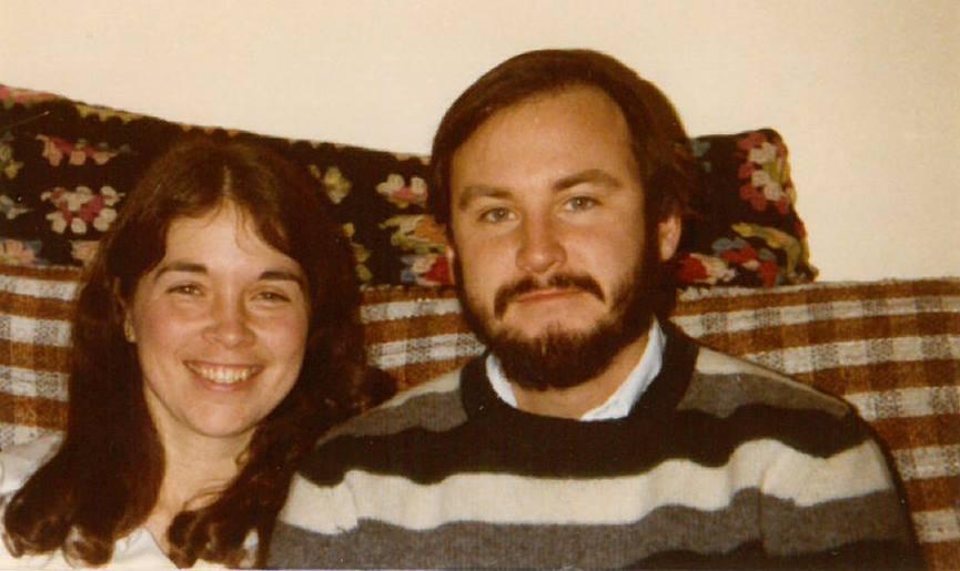 A smiling woman and bearded man sit together on a patterned sofa, exuding warmth and happiness. The image conveys a sense of comfort and togetherness.