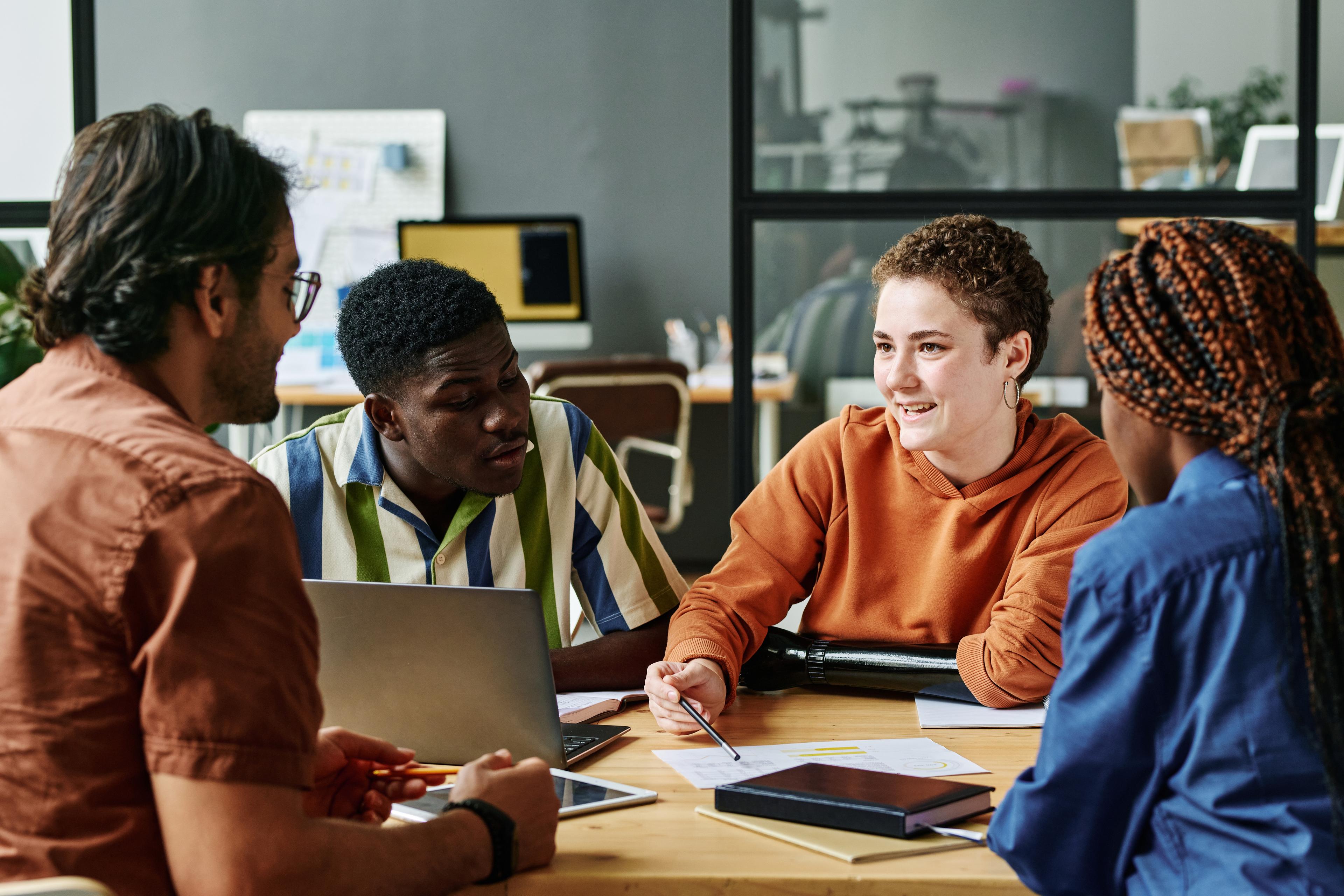 A diverse group of four people sit around a table in a bright office, engaged in a lively discussion. Laptops, notebooks, and papers are visible.