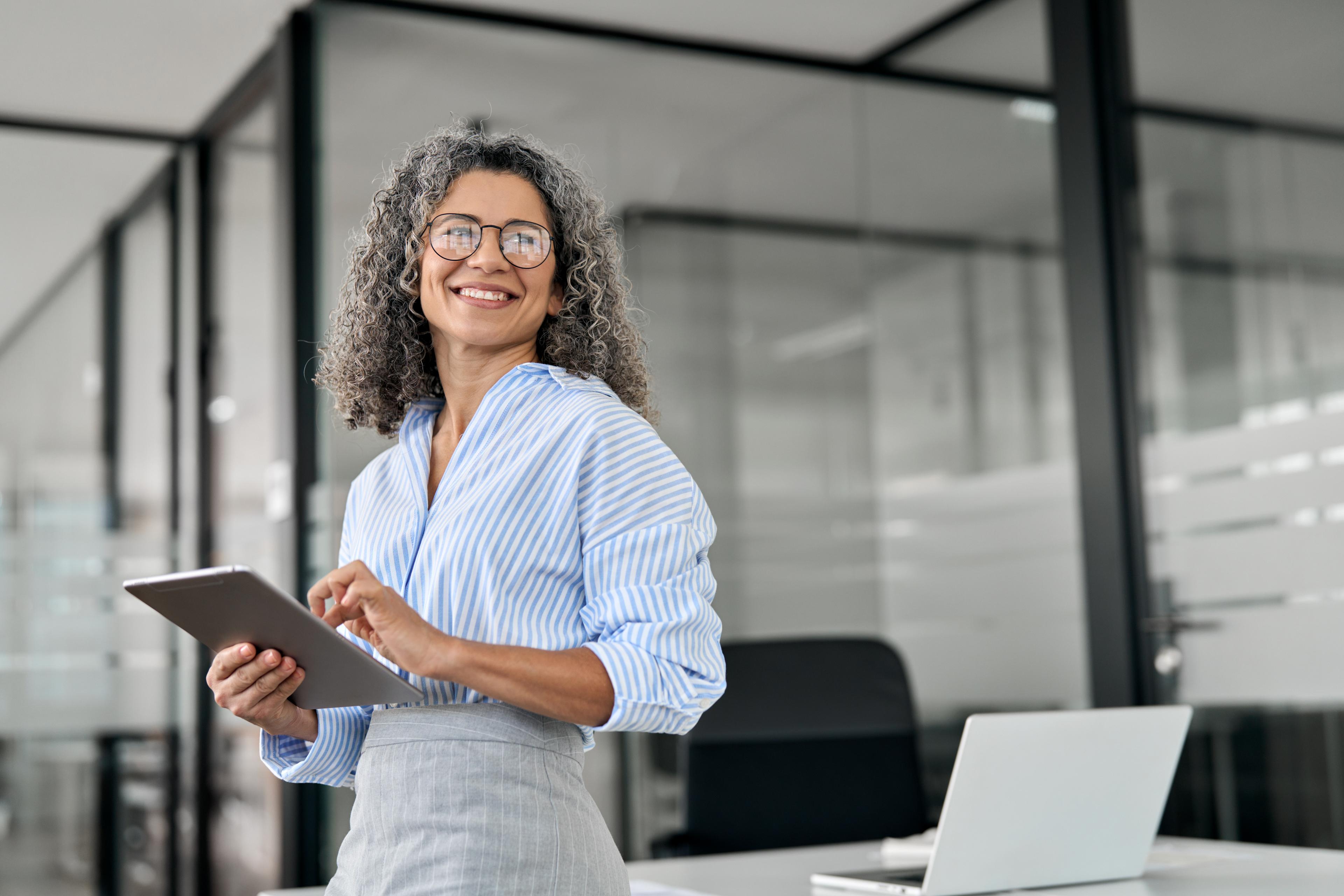 Smiling woman with curly gray hair and glasses stands in an office holding a tablet. She wears a blue-striped shirt and gray skirt, conveying confidence.